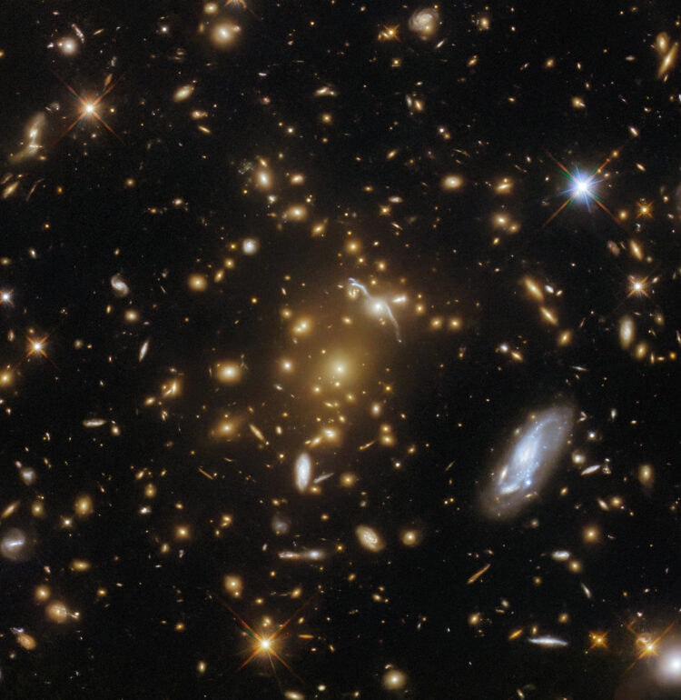 A vast galaxy cluster lurks in the centre of this image from the NASA/ESA Hubble Space Telescope. Like a submerged sea monster causing waves on the surface, this cosmic leviathan can be identified by the distortions in spacetime around it. The mass of the cluster has caused the images of background galaxies to be gravitationally lensed; the galaxy cluster has caused a sufficient curvature of spacetime to bend the path of light and cause background galaxies to appear distorted into streaks and arcs of light. A host of other galaxies can be seen surrounding the cluster, and a handful of foreground stars with tell-tale diffraction spikes are scattered throughout the image.This particular galaxy cluster is called eMACS J1823.1+7822, and lies almost nine billion light-years away in the constellation Draco. It is one of five exceptionally massive galaxy clusters explored by Hubble in the hopes of measuring the strengths of these gravitational lenses and providing insights into the distribution of dark matter in galaxy clusters. Strong gravitational lenses like eMACS J1823.1+7822 can help astronomers study distant galaxies by acting as vast natural telescopes which magnify objects that would otherwise be too faint or distant to resolve.