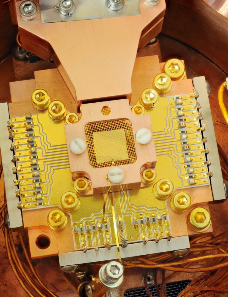 NIST physicists used this apparatus to coax two beryllium ions (electrically charged atoms) into swapping the smallest measurable units of energy back and forth, a technique that may simplify information processing in a quantum computer. The ions are trapped about 40 micrometers apart above the square gold chip in the center. The chip is surrounded by a copper enclosure and gold wire mesh to prevent buildup of static charge.