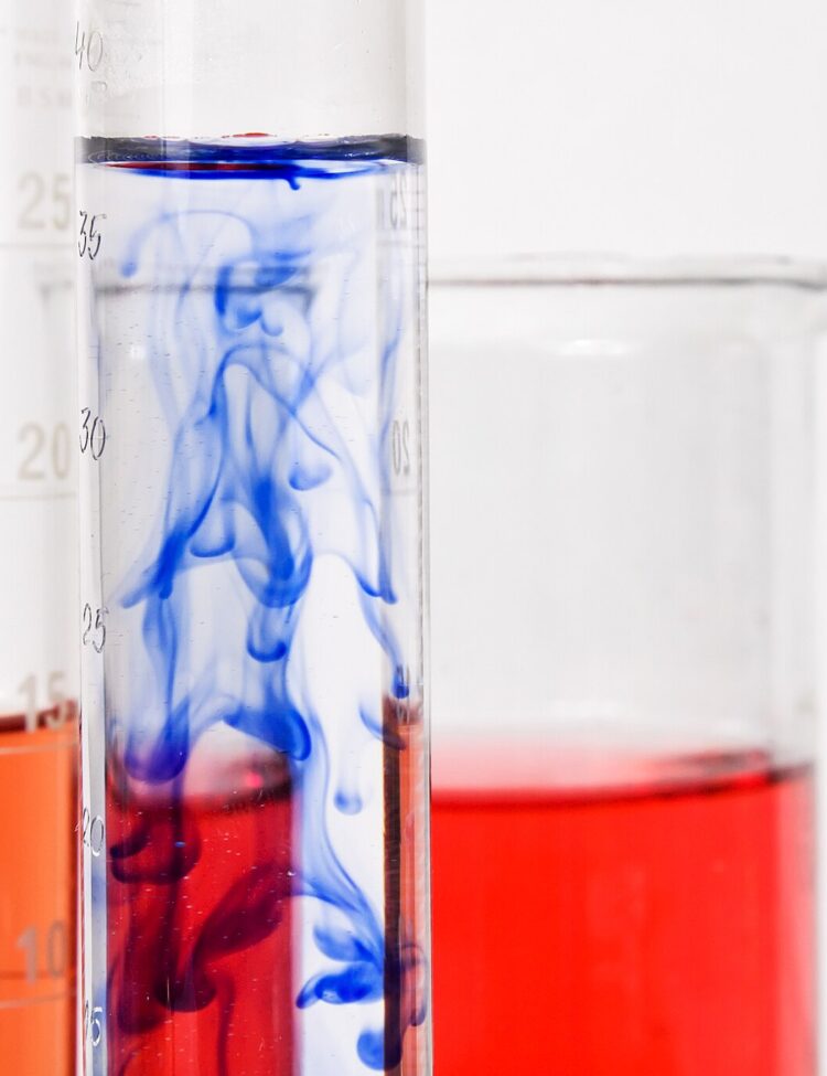 Several graduated cylinders of various thickness and heights with white side markings in front of a large beaker. They are all filled about halfway with red or blue chemical compounds. The blue ink is showing signs of Brownian motion when dissolving into water.