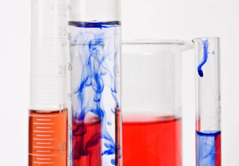 Several graduated cylinders of various thickness and heights with white side markings in front of a large beaker. They are all filled about halfway with red or blue chemical compounds. The blue ink is showing signs of Brownian motion when dissolving into water.