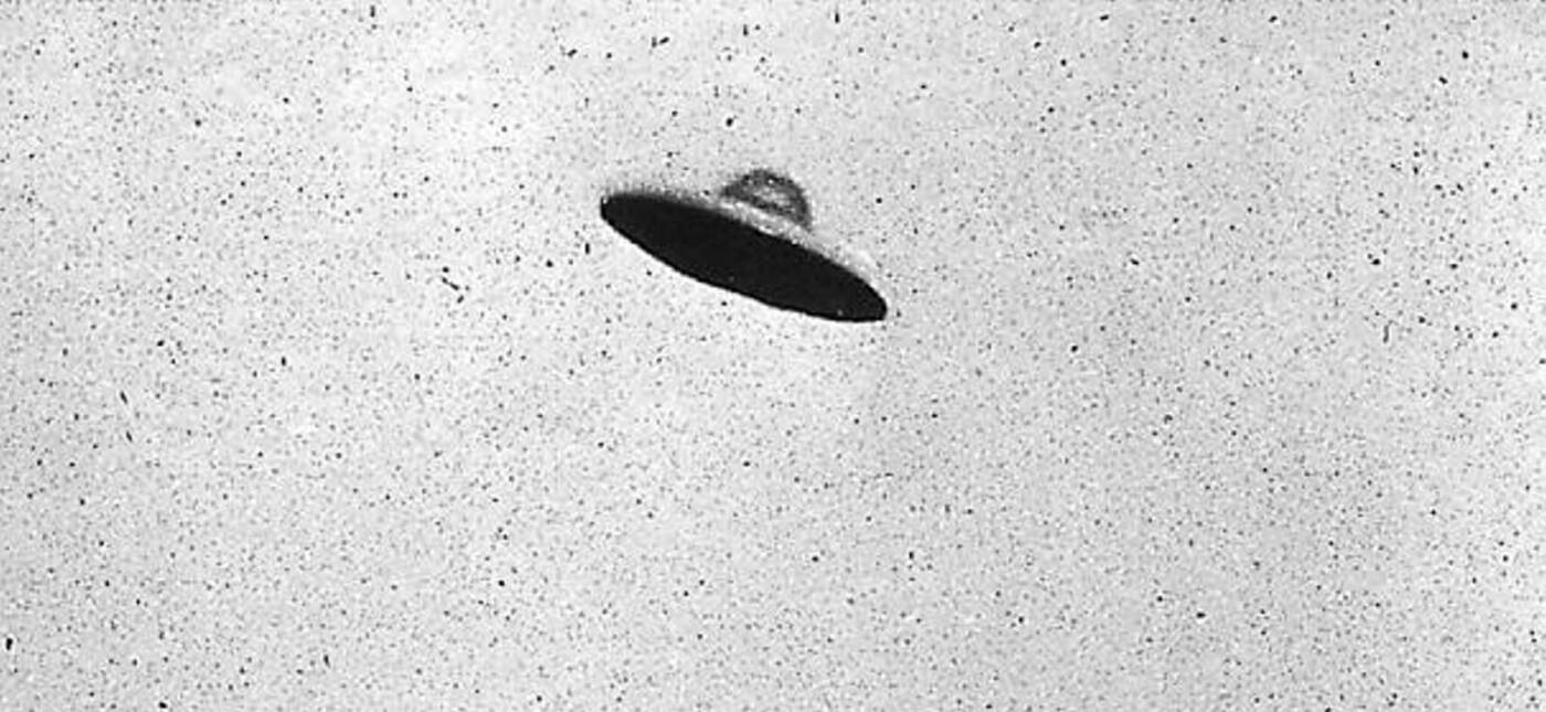 monotone non horizontal view of a supposed curvilinear centrally domed UFO relatively motionless (no blur) above an electrocommunications pole plus cables with deciduous trees.